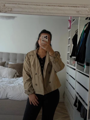 Kort beige trenchcoat med dubbelknäppning - Säljer en snygg kort trenchcoat i beige med klassisk dubbelknäppning och axelklaffar. Jackan har breda slag, knappar på ärmsluten och en avslappnad passform. Perfekt för lager-på-lager och stilrena outfits.