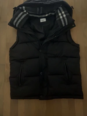 Burberry väst  - Snygg svart dunväst från Burberry med avtagbar huva och klassiskt rutigt foder. Västen har två dragkedjeförsedda fickor och knäppning framtill. Perfekt för lager-på-lager och håller dig varm under kyliga dagar.