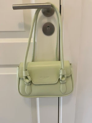Ljusgrön axelremsväska i konstläder - Trendig ljusgrön handväska med axelremmar och snygga spänndetaljer framtill. Väskan är tillverkad i konstläder och har en stilren, rektangulär form. Perfekt för dig som vill ha en poppig accessoar till din outfit.