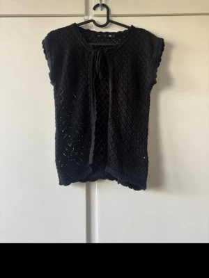 Svart stickad topp med knyt - Svart stickad topp med korta ärmar och knyt framtill. Toppen har ett diskret hålmönster och är lätt croppad. Perfekt för att skapa en chill och trendig look.