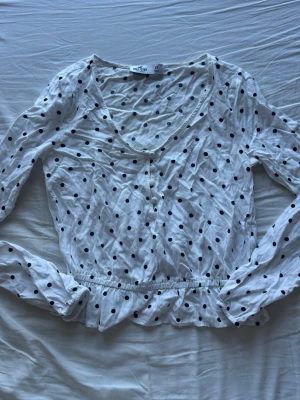 Vit prickig blus från Hollister - Supersöt vit blus från Hollister med svarta prickar, v-ringning och knappar framtill. Blusen har långa ärmar och resår i midjan som ger en snygg passform. 