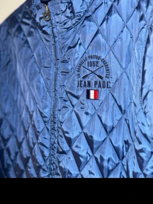 Blå quiltad jacka från Jean Paul - Snygg blå quiltad jacka från Jean Paul med dragkedja framtill och ståkrage. Jackan har ett broderat logotypmärke på bröstet och diskreta fickor i sidorna. Perfekt för dig som gillar stilren och sportig look.