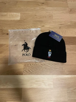 Svart mössa Polo Ralph Lauren - Säljer en svart ribbstickad mössa från Polo Ralph Lauren med den klassiska björnen broderad framtill. Mössan har uppvikt kant och är tillverkad i mjukt material som håller dig varm. Perfekt accessoar för kyliga dagar.