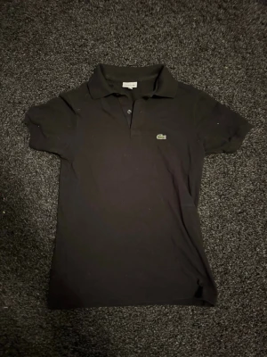 Svart pikétröja  Lacoste - Klassisk svart pikétröja från Lacoste med den ikoniska krokodilloggan på bröstet. Tröjan har krage, knappar framtill och korta ärmar. Perfekt till jeans och shorts  för en clean och sportig look.