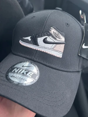 Svart Nike keps med sneakerbrodyr - Svart snapback-keps från Nike med broderad sneaker (Air Jordan 1) på framsidan. Justerbar passform med metallspänne bak och Nike-logga på insidan. Kepsen är i klassisk form och tillverkad i bomullstyg. Skriv gärna för frågor!