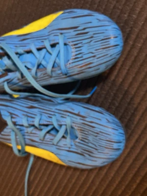 Blå fotbollsskor från Puma - Säljer ett par blå fotbollsskor från Puma med gula detaljer och svarta ränder. Skorna har snörning och dobbar i plast för bra grepp på planen. Snygg design med Pumas logga på sidan och en sportig look som sticker ut på matchen.