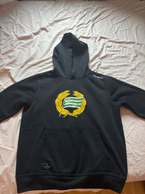 Svart Hammarby hoodie från Craft - Svart hoodie från Craft med Hammarby IF:s emblem i gult och grönt på bröstet. Klassisk huva och stor magficka framtill. Perfekt för dig som vill visa ditt stöd för Hammarby och samtidigt vara bekväm. Mjuk bomullsblandning.