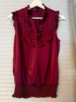 Vinröd blus med volangdetaljer - Snygg vinröd blus med volangkantad krage och knappar framtill. Ärmlös modell med smockad nederkant och dekorativa veck på bröstet. Perfekt för dig som gillar en feminin och trendig stil.