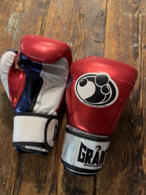 Röd och vit boxningshandskar Grant - Säljer ett par boxningshandskar från Grant i rött, vitt, blått och silver. Handskarna har en snygg metallic finish och tydliga loggor på ovansidan och vid handleden. Perfekta för träning eller sparring inom boxning.