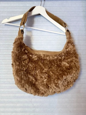 Brun fluffig axelväska med bred rem - Supermjuk och fluffig handväska i brun fuskpäls med bred axelrem i beige konstläder och snygga guldfärgade detaljer. Väskan har en rundad form och stängs med dragkedja upptill. Perfekt för dig som vill ha en trendig accessoar med cozy vibe.