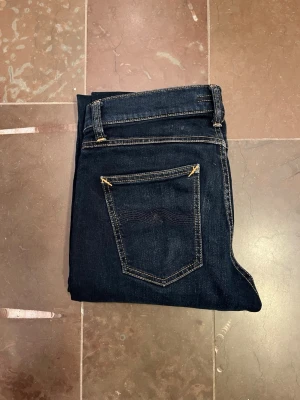 Mörkblå jeans från Nudie Jeans - Säljer ett par mörkblå jeans från Nudie Jeans med klassisk femficksmodell och kontrastsömmar. Jeansen har smal passform och är tillverkade i mjukt denimtyg. Perfekta för dig som gillar stilrena och tidlösa jeans.