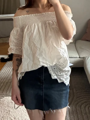 Vit offshoulder blus med spetsdetaljer - Söt vit offshoulder blus med broderade spetsdetaljer och korta ärmar. Blusen har en smockad överdel och luftig passform, perfekt för varma dagar. Romantisk och trendig look som passar till både jeans och kjol.