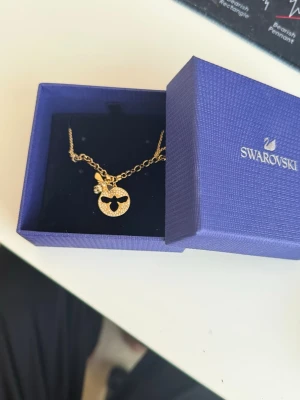 Guldigt halsband med bi från Swarovski - Unikt halsband från Swarovski i guldfärgad metall med ett runt hänge dekorerat med gnistrande stenar och en detalj i form av en liten bi. Kedjan har sexkantiga länkar som ger ett coolt och trendigt intryck. Perfekt för dig som gillar statement-smycken och vill sticka ut.