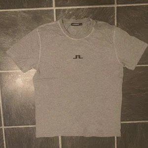 Grå t-shirt från J.Lindeberg -  Grå t-shirt från J.Lindeberg med svart logga på bröstet i storlek M