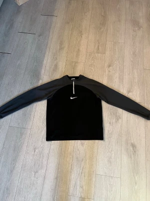 Nike xs  - Sportig svart långärmad topp från Nike med vit logga på bröstet och half zip-dragkedja framtill. Tillverkad i ett mjukt syntetmaterial som passar perfekt för träning eller en aktiv vardag. Enkel och stilren design med normal passform.