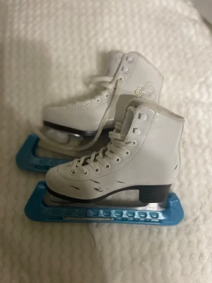 Vita konståkningsskridskor Spin - Vita konståkningsskridskor från Spin med högt skaft, snörning och blåa skydd på skenorna. Klassisk design med svart sula och detaljer i metall. Perfekta för isbanan eller träning. Har repor på sidan