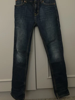 Mörkblå Levi's jeans med slitningar storlek 14A - Säljer ett par mörkblå Levi's jeans i storlek 14 A med snygga slitningar framtill och klassiska kontrastsömmar. Jeansen har slim fit passform, fem fickor och den ikoniska röda Levi's-lappen bak. Tillverkade i slitstark denim som ger en tidlös look. Hör av er vid minsta fråga eller funderingar 🙏 ej prut bart!