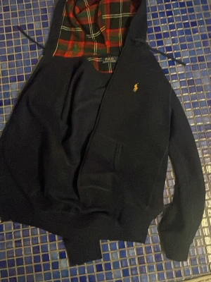Marinblå hoodie från Polo Ralph Lauren - Jag säljer min Polo Ralph Lauren tröja då jag aldrig använt den eftersom att den är för liten. Jag köpte tröjan och hängde direkt upp den i mitt skåp för att sen aldrig använda😓Det är en snygg marinblå hoodie från Polo Ralph Lauren med klassisk gul logga på bröstet. Insidan av huvan har ett rött och grönt rutigt foder som ger en unik touch. Hoodien har känguruficka och ribbade muddar. Perfekt för en avslappnad och stilren look.