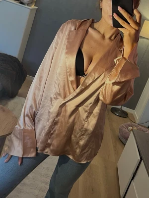 Champagnefärgad/ rosa satinblus  - Säljer en champagnefärgad satinblus från Gina Tricot med avslappnad passform och breda manschetter. Blusen har djup v-ringning, krage och långa ärmar. Perfekt att styla med jeans eller kjol för en trendig look.