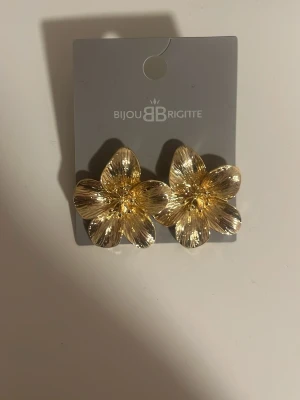 Guldiga blomörhängen Bijou Brigitte - Stora, blommiga örhängen i guldfärgad metall från Bijou Brigitte. Örhängena har en elegant och glansig finish med detaljerade blomblad som ger ett lyxigt intryck. Perfekta för dig som vill sticka ut med accessoarer som verkligen syns.