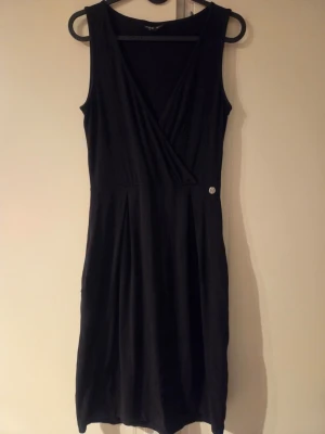 Svart ärmlös omlottklänning - Stilren svart ärmlös klänning med omlottöverdel och markerad midja. Klänningen har en liten dekorativ knapp i midjan och faller i knälång längd. Tillverkad i mjukt, stretchigt tyg som ger en bekväm passform. Perfekt för dig som gillar enkel och klassisk stil. Storlek XS, men mer som S.