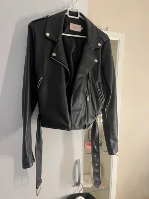 Svart skinnjacka från Nelly - Snygg svart skinnjacka från Nelly med klassisk bikerstil, silvriga dragkedjor och knappar samt bälte i midjan. Jackan har breda slag och två snedställda fickor med dragkedja. Perfekt för en cool och trendig look.