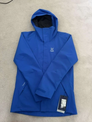 Blå regnjacka från Haglöfs - Snygg blå regnjacka från Haglöfs med huva och dragkedja framtill. Jackan har två sidofickor med dragkedja och justerbara ärmslut med kardborre. Tillverkad i slitstarkt, vattenavvisande material som passar perfekt för regniga dagar. Diskret Haglöfs-logga på bröstet.