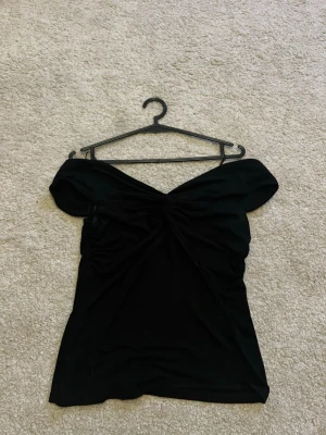 Svart offshoulder topp - Snygg svart offshoulder topp med twistad detalj framtill. Toppen har en figurnära passform och är perfekt för att lyfta vilken outfit som helst. Tillverkad i mjukt material som sitter skönt mot kroppen.