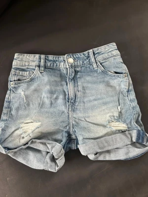 Blå denimshorts från H&M - Säljer ett par blå denimshorts från H&M med avslappnad passform. Shortsen har slitna detaljer, uppvikta benslut och klassisk femficksdesign. Perfekta för varma dagar och ger en cool, avslappnad look.