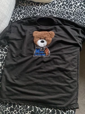 Svart t-shirt med nalle från Made Extreme - Svart t-shirt från Made Extreme med ett stort broderat nallehuvud i brunt och vitt på bröstet. Under nallen finns en blå och orange logga. T-shirten har klassisk passform och är kortärmad, perfekt för en cool streetwear-look.