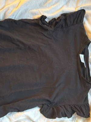 Svart volangtopp från Gina Tricot - Svart topp från Gina Tricot med volangdetaljer vid axlarna. T-shirten är i mjuk bomull och har en klassisk rund halsringning. Perfekt för dig som gillar en enkel men ändå söt stil.