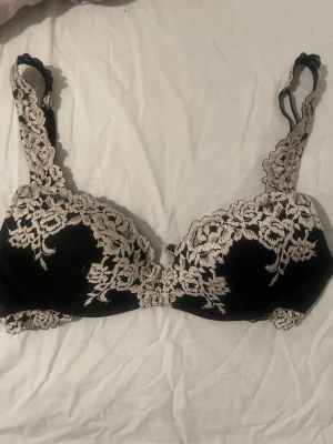 Svart spets-bh med beige brodyr Superpushup-behå Gioia Pretty Flowers - Snygg svart bh med vadderade kupor och vacker beige spetsbrodyr över hela framsidan och axelbanden. Bh:n har bygel och justerbara axelband för bästa passform. Perfekt för dig som gillar en feminin och elegant stil.