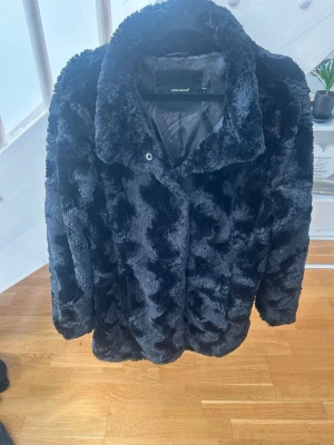 Svart fuskpälsjacka Vero Moda - Snygg svart fuskpälsjacka från Vero Moda med bred krage och dold knäppning framtill. Jackan har en mjuk och fluffig yta som ger en lyxig känsla och är helfodrad för extra värme. Perfekt för kalla vinterdagar och ger en trendig look.