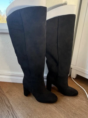 Svarta höga stövlar med klack - Säljer ett par svarta höga stövlar i mockaimitation med bred klack och rundad tå. Stövlarna går upp över knät och har en stilren design utan synliga detaljer eller dragkedjor. Perfekta för att lyfta vilken outfit som helst.