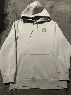 Ljusgrå hoodie med tryck - Ljusgrå hoodie med huva och dragsnören, samt en liten grafisk detalj på bröstet. Klassisk känguruficka framtill och ribbade muddar vid ärmslut och nederkant. Mjuk bomullskänsla och stilren design som passar till det mesta.