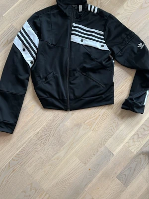 Svart Adidas track jacket DAM - Snygg svart bomberjacka från adidas med vita och grå ränder på axlar och bröst. Jackan har dragkedja framtill, två fickor och adidas logga broderad på ärmen. Tillverkad i polyester och har en croppad passform som ger en sportig vibe.