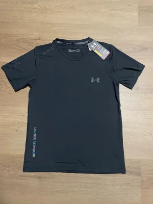 Svart tränings-tshirt från Under Armour - Snygg svart tränings-tshirt från Under Armour med UA-logga på bröstet och diskret text på axeln. Tillverkad i lätt och snabbtorkande material, perfekt för löpning eller gym. Rund halsringning och korta ärmar. Under Armour-tryck längs sidan.