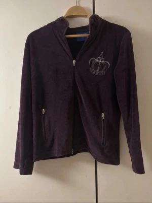 Lila velour hoodie med krona - Mysig lila hoodie i velour från morgensteen med dragkedja och två fickor framtill. Broderad krona på bröstet och ryggen ger en lyxig vibe. Perfekt för dig som gillar en avslappnad men ändå trendig stil.