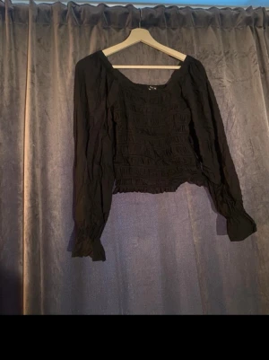 Svart croppad blus off shoulder - Trendig svart croppad blus med långa puffärmar och smockdetaljer över hela toppen. Fyrkantig ringning och elastiska muddar vid ärmsluten ger en snygg siluett. Perfekt att styla med höga jeans eller kjol.