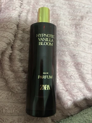 Zara Hypnotic Vanilla Bloom parfym - En elegant parfym från Zara med namnet Hypnotic Vanilla Bloom. Flaskan är cylinderformad i mörklila glas med guldfärgat lock och text i guld. Doften har en tydlig vaniljkaraktär och flaskan känns lyxig och modern – perfekt för dig som gillar söta och varma toner.