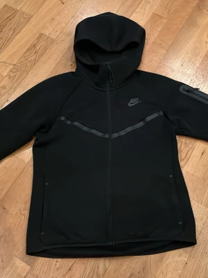 Svart Nike Tech Fleece hoodie - Svart Nike Tech Fleece hoodie med hel dragkedja, huva och diskret Nike-logga på bröstet. Snygga svarta detaljer över bröstet och en praktisk ficka med dragkedja på ärmen. Perfekt för en sportig och stilren look.