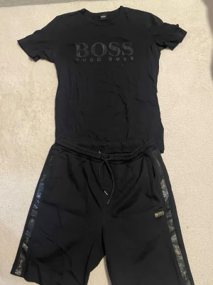 Svart set från Hugo Boss - Snyggt svart set från Hugo Boss med t-shirt och shorts. T-shirten har diskret BOSS-logga framtill och rund hals. Shortsen har dragsko i midjan, sidofickor och BOSS-detaljer längs sidorna. Perfekt för en avslappnad och stilren look.