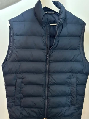 Mörkblå dunväst från Zara - Snygg svart dunväst från Zara med quiltad design och dragkedja framtill. Västen har två sidofickor och hög krage som ger en cool look. Perfekt att slänga över en hoodie för extra värme. Materialet är lätt och puffigt.