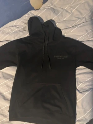 Svart Essentials Fear of God hoodie - Säljer en svart hoodie från Essentials Fear of God med diskret logga på bröstet och ärmen. Hoodien har stor huva, dragsnören och en klassisk magficka. Tillverkad i mjukt material som passar perfekt för en avslappnad streetwear-look.