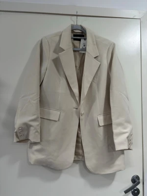 Beige kavaj  - Stilren beige kavaj med klassisk passform och breda slag. Kavajen har två fickor med lock framtill och knäppning med en knapp. Ärmarna har dekorativa knappar vid mudden. Perfekt för att höja din outfit.