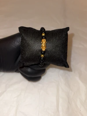 Svart pärlarmband med guldfärgad detalj - Snyggt armband med svarta pärlor och guldfärgade detaljer, inklusive en liten figur i mitten. Armbandet har en stilren och modern look som passar perfekt till olika outfits. Elastiskt band för enkel påtagning.