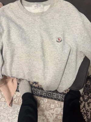 Grå sweatshirt från Moncler - Säljer en stilren grå sweatshirt från Moncler med rund halsringning och diskret Moncler-logga broderad på bröstet. Tröjan har långa ärmar och ribbade muddar vid ärmslut och nederkant. Perfekt för en avslappnad och trendig look.