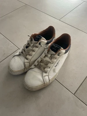 Vita sneakers från Pepe Jeans - Snygga vita sneakers från Pepe Jeans med bruna och mörkblå detaljer. Skorna har lågt skaft, vita snören och logga broderad på sidan. Tillverkade i läderimitation med textilfoder och gummisula. Perfekta för en avslappnad och trendig stil.