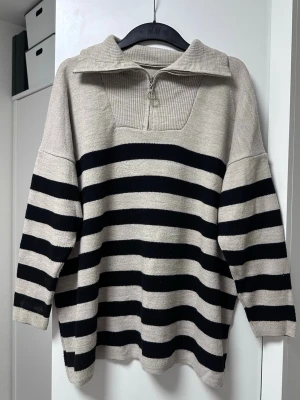 Beige och svart randig stickad tröja - Stickad tröja från H&M i beige med svarta horisontella ränder. Tröjan har en bred krage med halv dragkedja och ribbstickad detalj vid halsen. Långärmad och avslappnad passform, perfekt för en chill och trendig look.