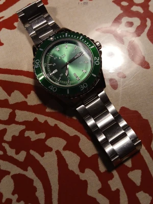 Grön klocka med metallarmband från Regal - Priset kan diskuteras. Snygg klocka från Regal med grön urtavla och matchande grön bezel. Klockan har ett robust metallarmband i silverfärg och tydliga markeringar på urtavlan. Perfekt accessoar för dig som vill sticka ut med stilren design.